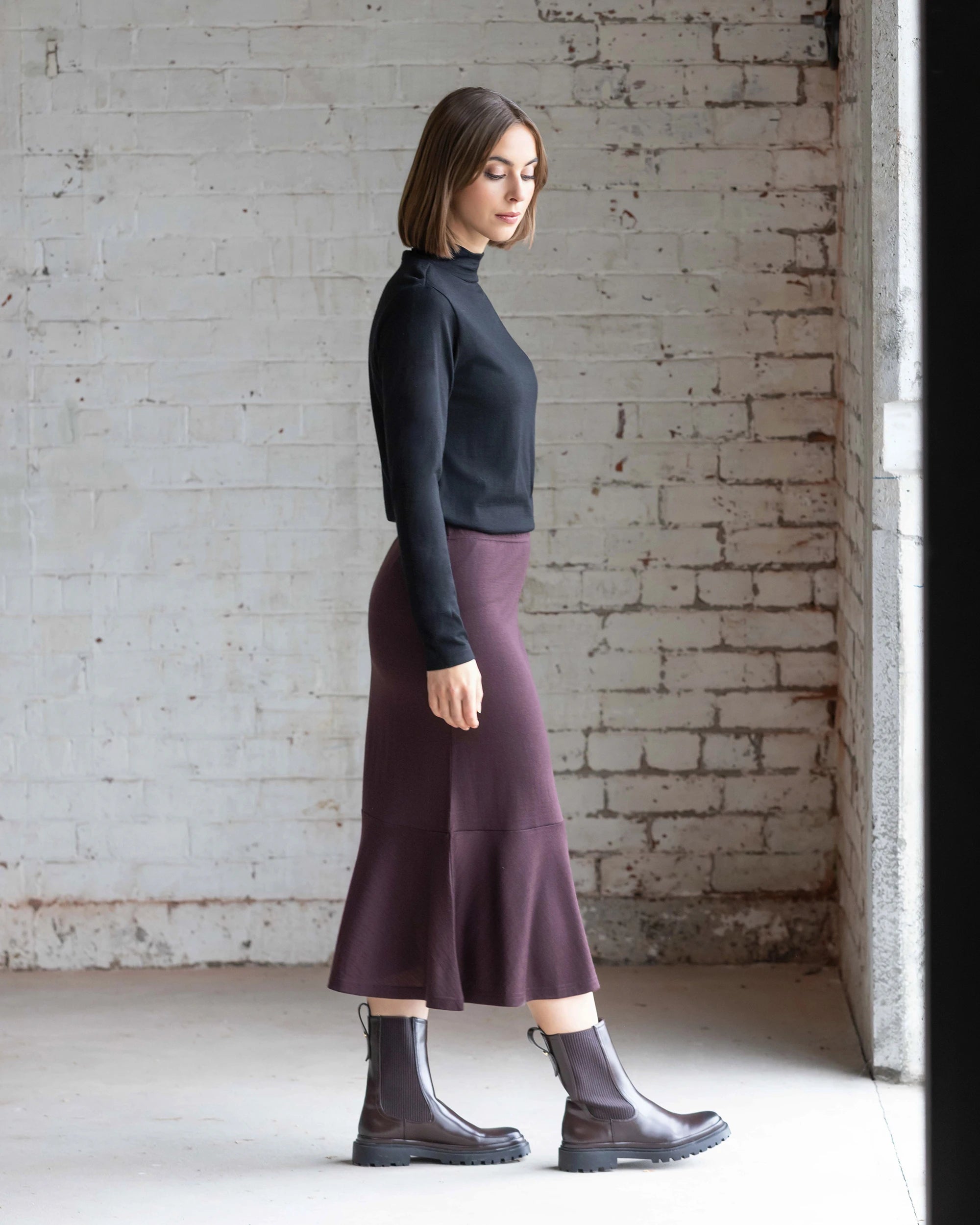 A woman with a bob stands indoors by a white brick wall, wearing a black long-sleeve top, the Vassalli Carrie Merino Skirt - Berry, and brown ankle boots, looking downward with her arms at her sides.