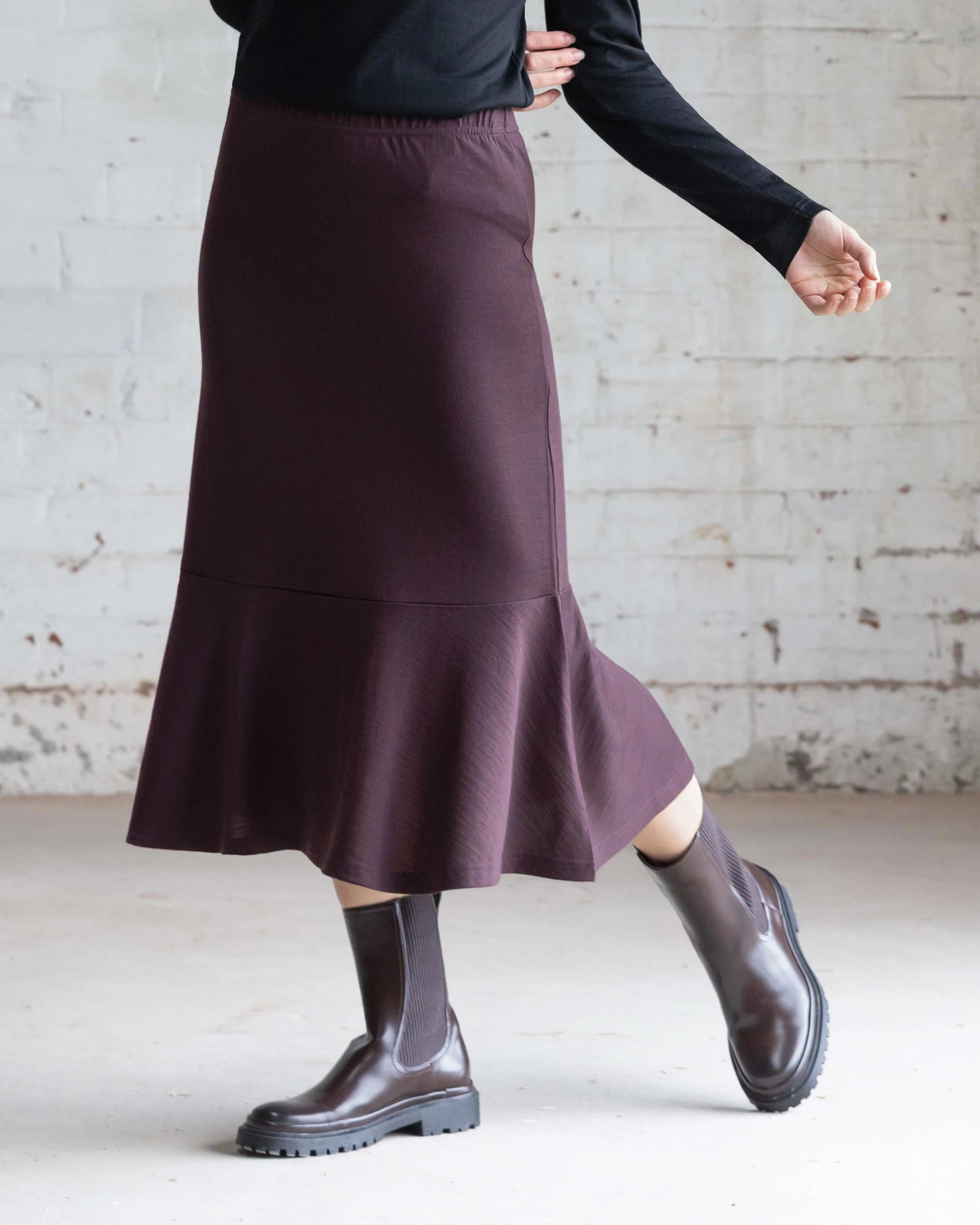 A person wearing the Vassalli Carrie Merino Skirt in Berry, a black top, and brown ankle boots stands against a white brick wall, with one arm bent and one foot slightly raised.