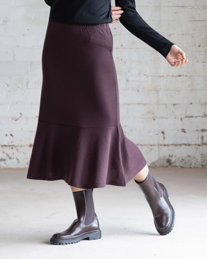 A person wearing the Vassalli Carrie Merino Skirt in Berry, a black top, and brown ankle boots stands against a white brick wall, with one arm bent and one foot slightly raised.