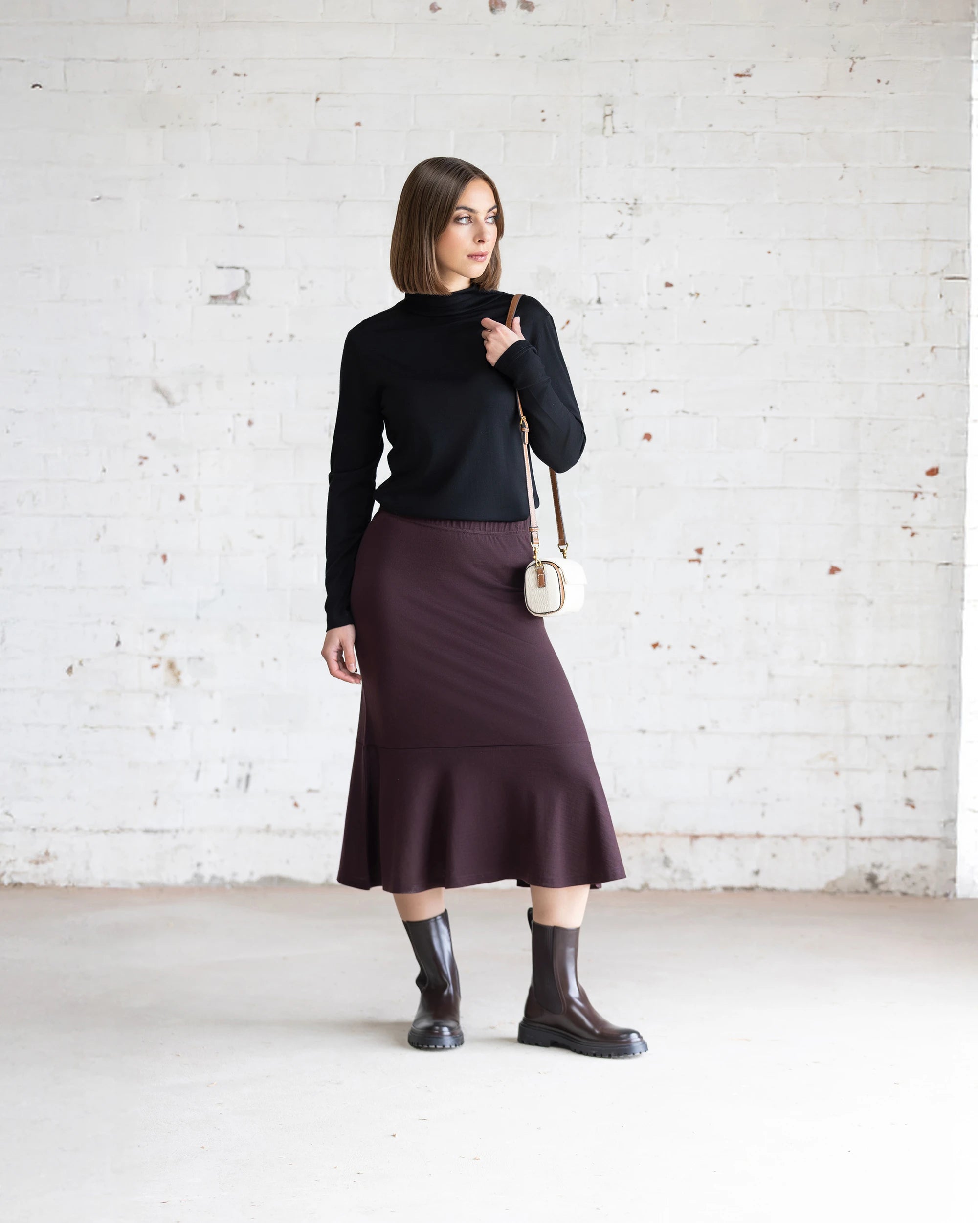 A woman stands in a minimalist room with a white brick wall, wearing the Vassalli Carrie Merino Skirt in Berry, paired with a black turtleneck, black boots, and a small white shoulder bag, looking to the side with one hand on her shoulder.