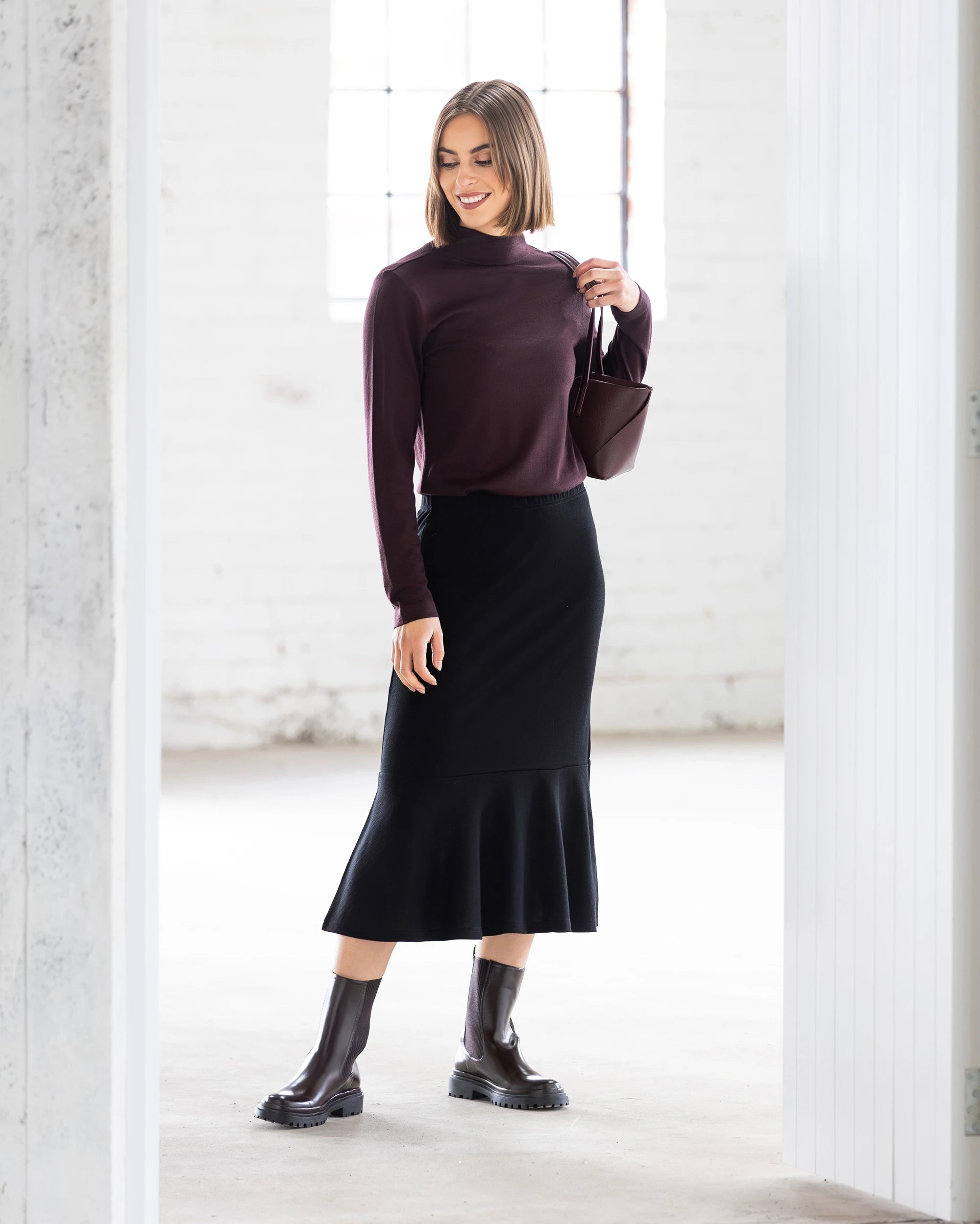 A woman smiles and looks to the side in a bright, minimalist room, wearing the Vassalli Carrie Merino Skirt in black, paired with a maroon long-sleeve top, dark ankle boots, and holding a small matching handbag.