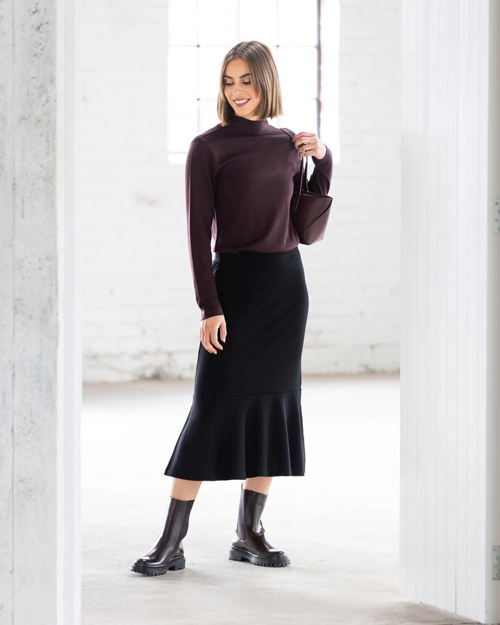 A woman smiles and looks to the side in a bright, minimalist room, wearing the Vassalli Carrie Merino Skirt in black, paired with a maroon long-sleeve top, dark ankle boots, and holding a small matching handbag.