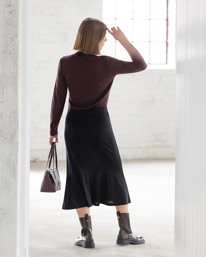 A woman with shoulder-length hair stands indoors facing away, wearing a burgundy top, brown boots, a matching handbag, and the Vassalli Carrie Merino Skirt in black with a peplum hem. She gestures near her head with her right hand.