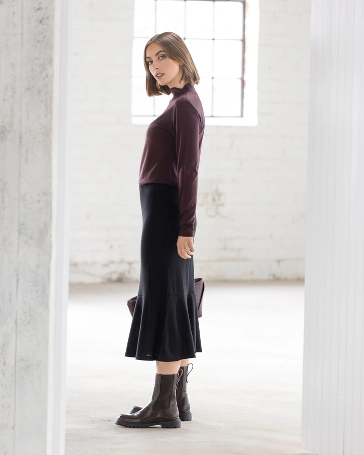 A woman with straight, chin-length hair stands sideways in a bright room, wearing a burgundy long-sleeve top, brown lace-up boots, and the Vassalli Carrie Merino Skirt in Black. She looks over her shoulder.