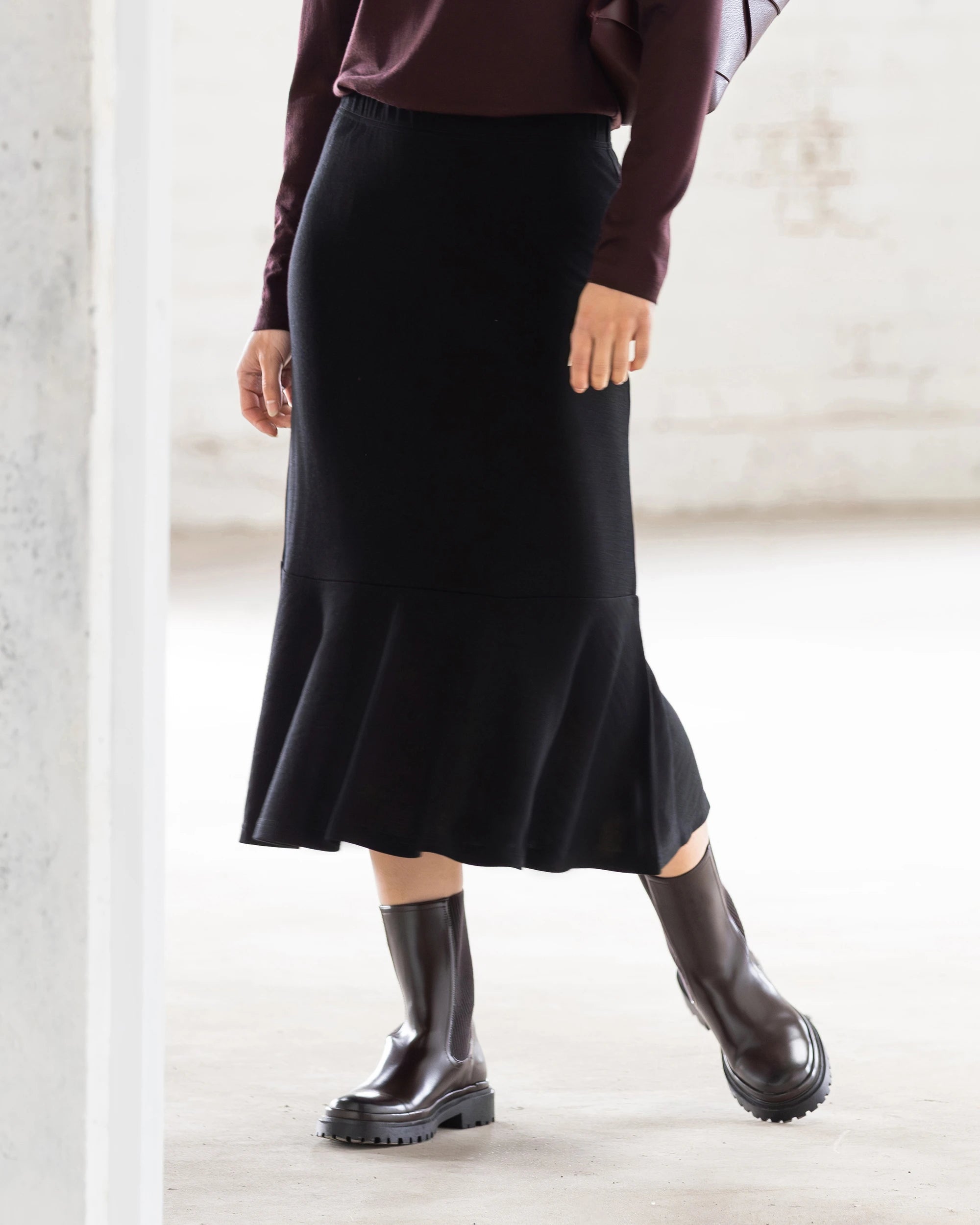 A person stands indoors with one knee slightly bent, wearing the Vassalli Carrie Merino Skirt in black, a dark long-sleeve top, and black ankle boots. The softly lit background is minimal.