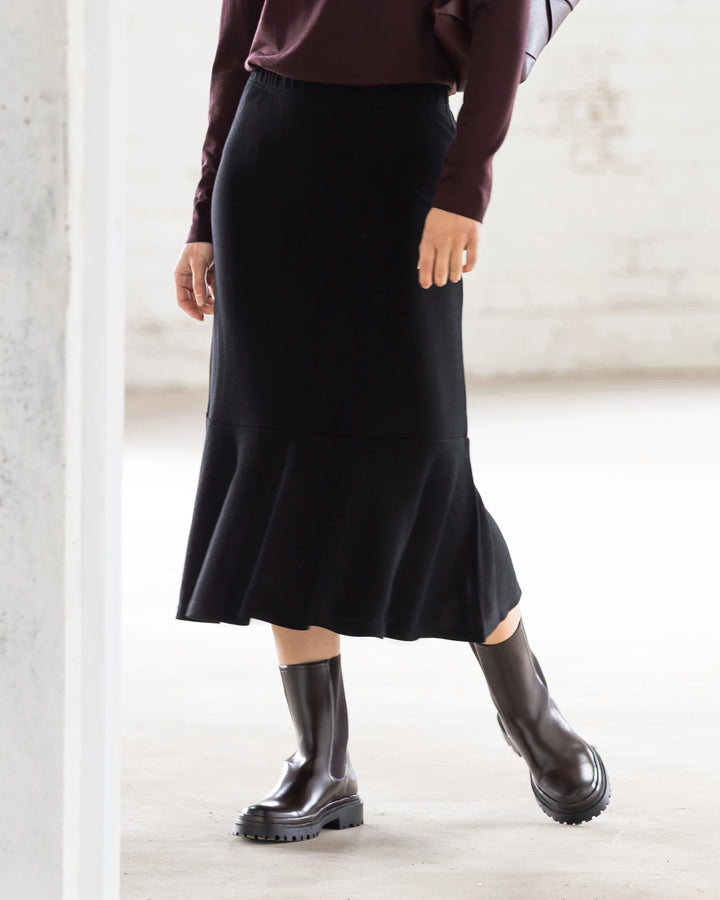 A person stands indoors with one knee slightly bent, wearing the Vassalli Carrie Merino Skirt in black, a dark long-sleeve top, and black ankle boots. The softly lit background is minimal.