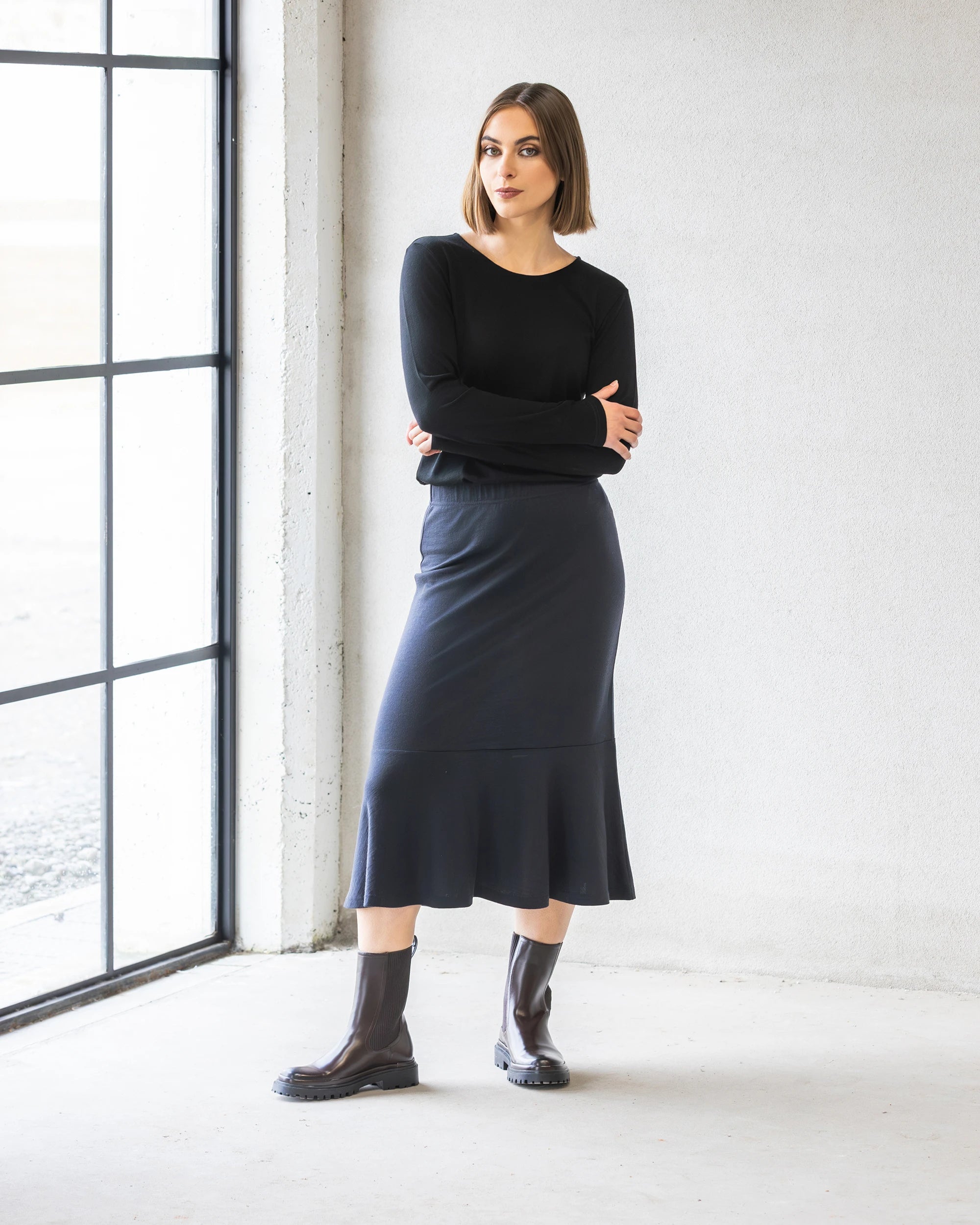 A woman with a straight bob poses indoors near a large window, arms crossed, wearing the Vassalli Carrie Merino Skirt in Midnight (midi-length), a black long-sleeve top, and dark brown ankle boots.