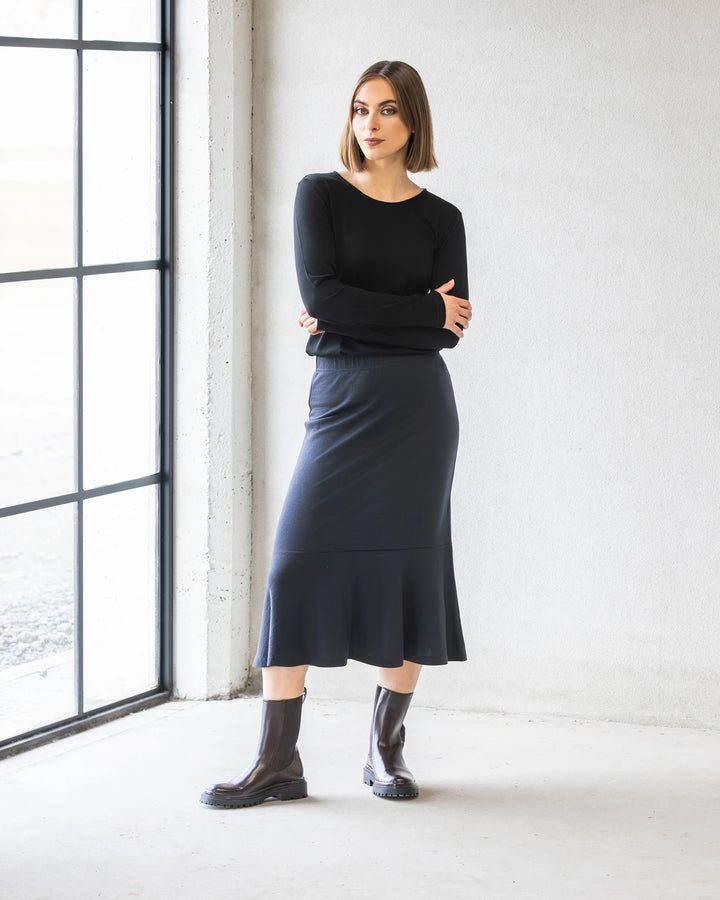 A woman with a straight bob poses indoors near a large window, arms crossed, wearing the Vassalli Carrie Merino Skirt in Midnight (midi-length), a black long-sleeve top, and dark brown ankle boots.