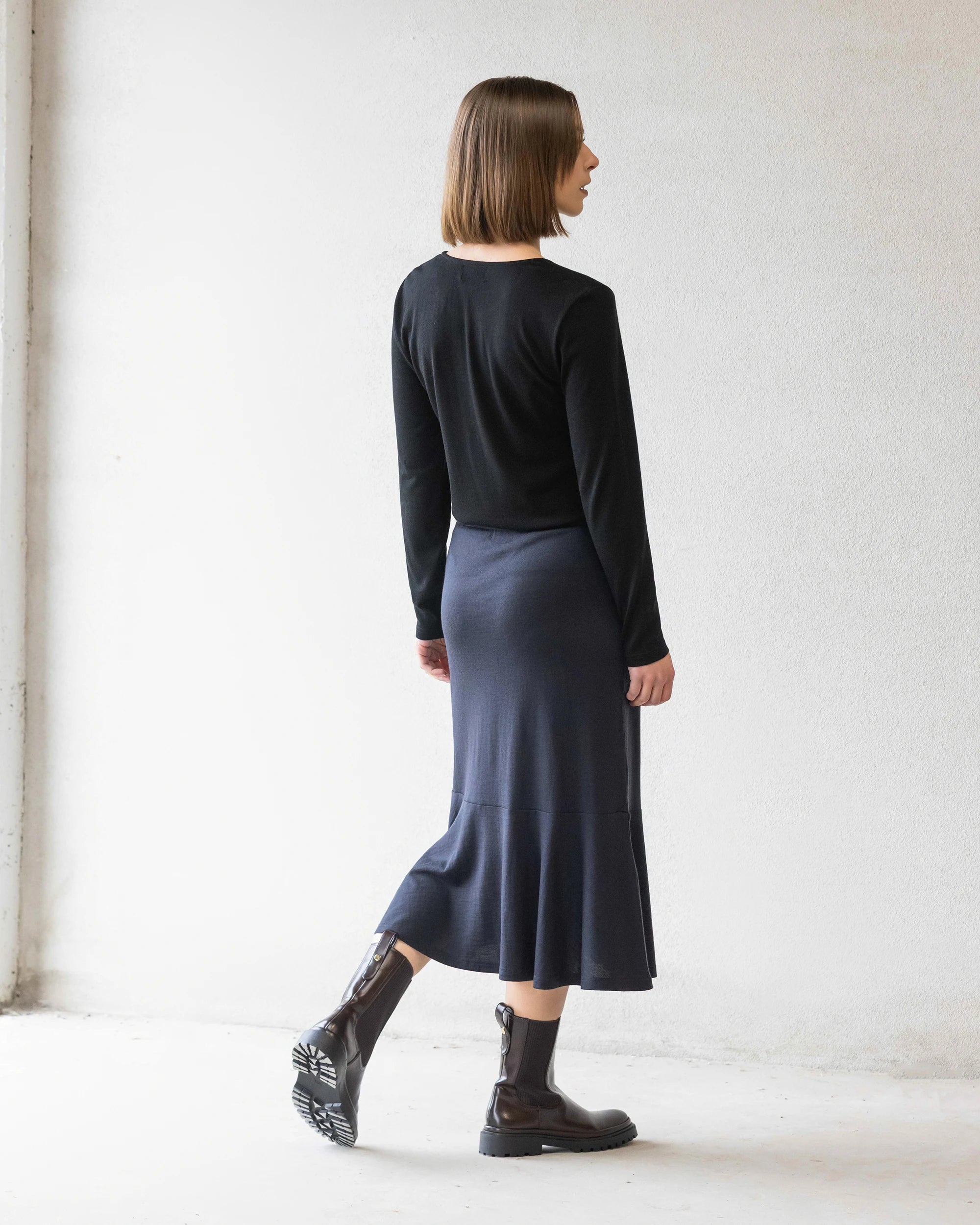 A woman with a straight bob stands facing away against a white wall, wearing a black long-sleeve top, black ankle boots, and the Vassalli Carrie Merino Skirt in Midnight.