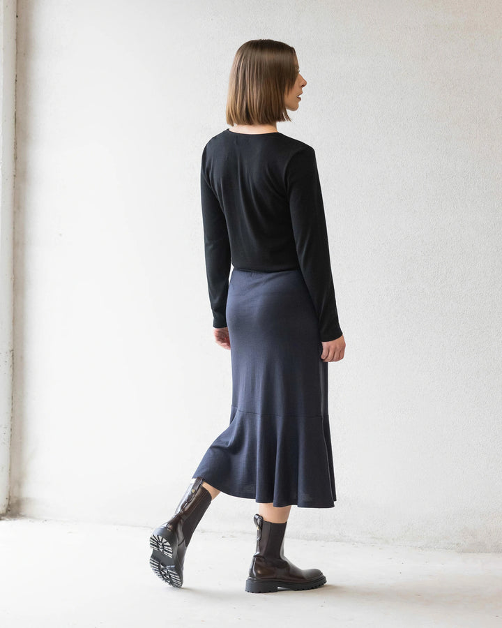A woman with a straight bob stands facing away against a white wall, wearing a black long-sleeve top, black ankle boots, and the Vassalli Carrie Merino Skirt in Midnight.