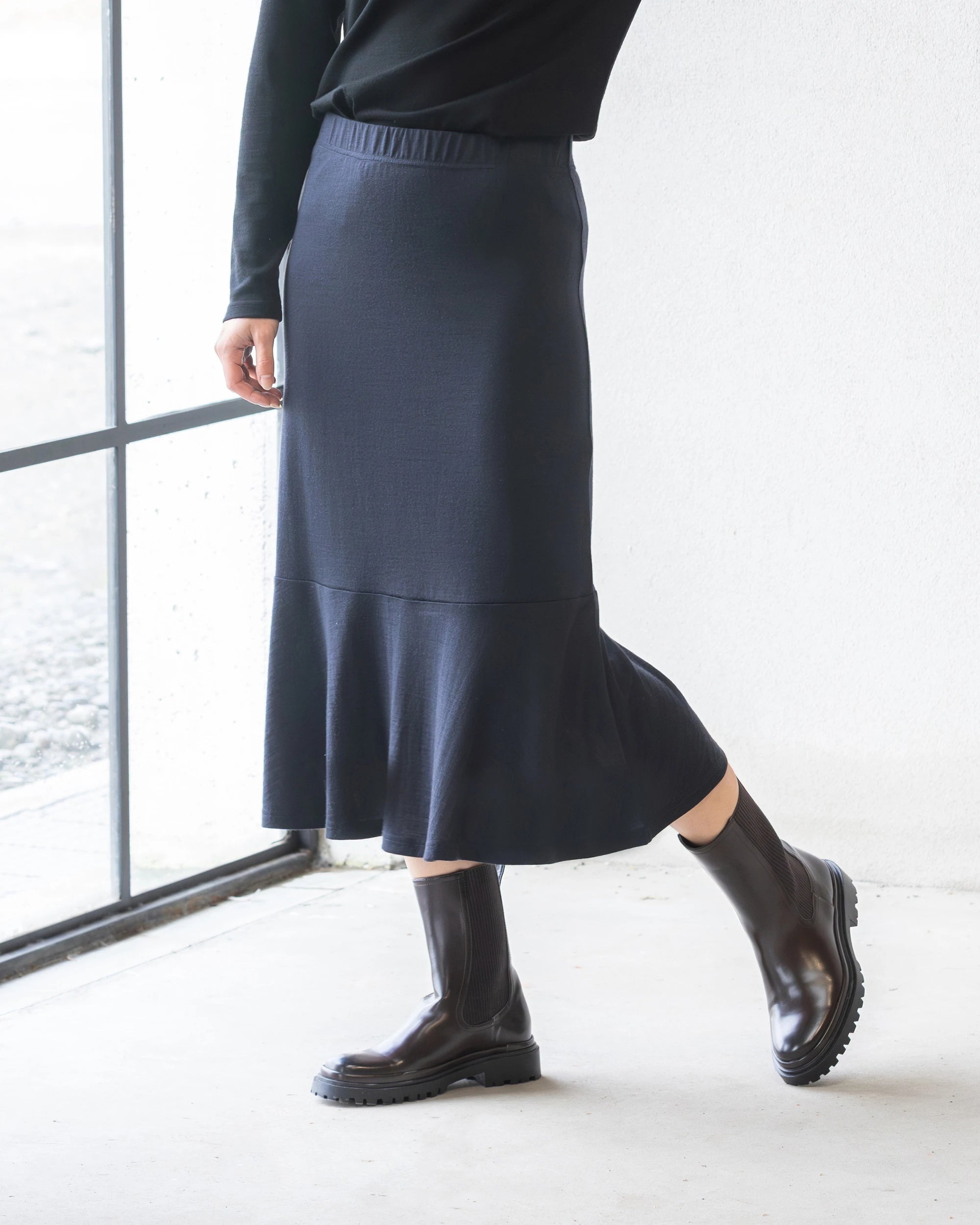 A person wearing a black long-sleeve top, the Vassalli Carrie Merino Skirt in Midnight with a flared peplum hem, and shiny black ankle boots stands indoors by a window, one foot slightly raised.