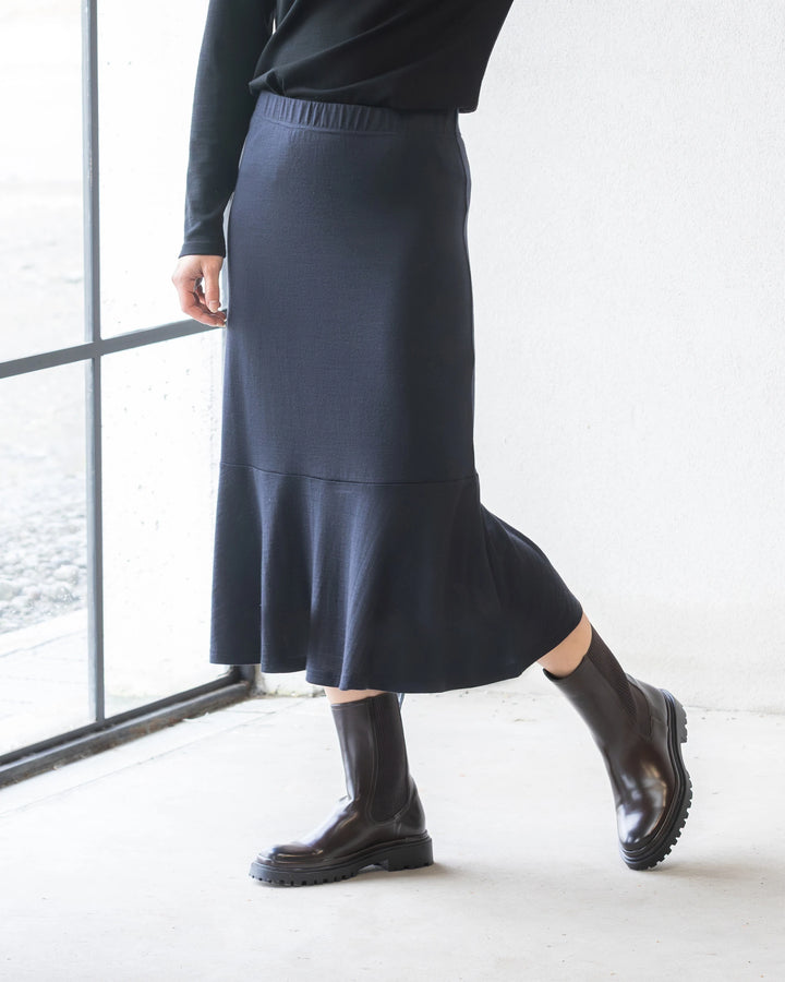 A person wearing a black long-sleeve top, the Vassalli Carrie Merino Skirt in Midnight with a flared peplum hem, and shiny black ankle boots stands indoors by a window, one foot slightly raised.