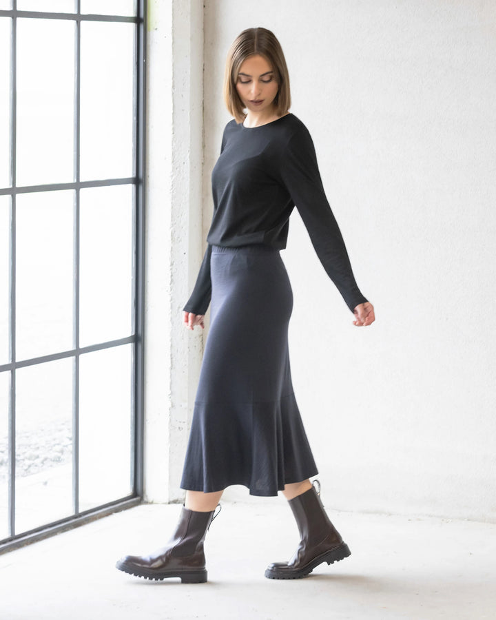 A woman with straight brown hair in a black long-sleeve top, the Vassalli Carrie Merino Skirt - Midnight, and dark brown chunky boots stands by a large window in a minimalist, light-filled room.