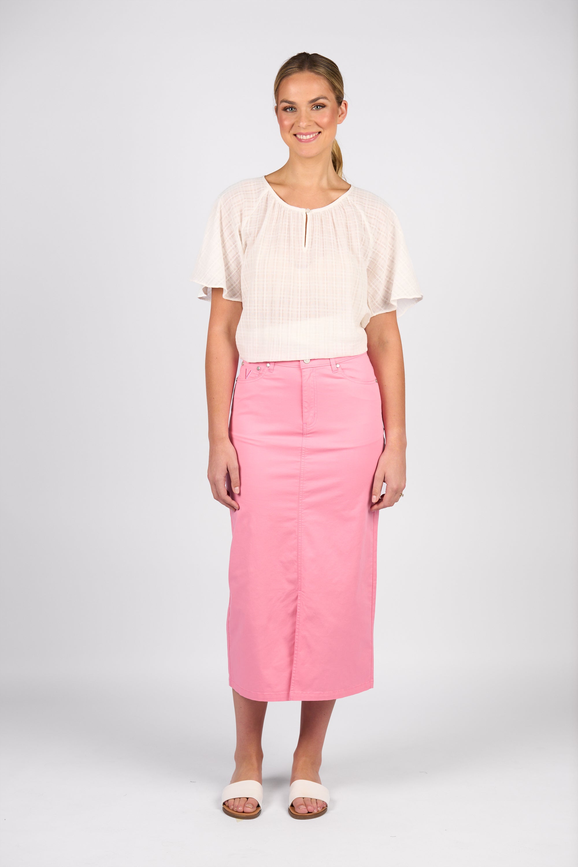 A woman smiles against a plain background, wearing a short-sleeved white blouse and the Vassalli Mid Length Skirt - Jam with white slide sandals. Her pulled-back hair completes her fresh, modern style.