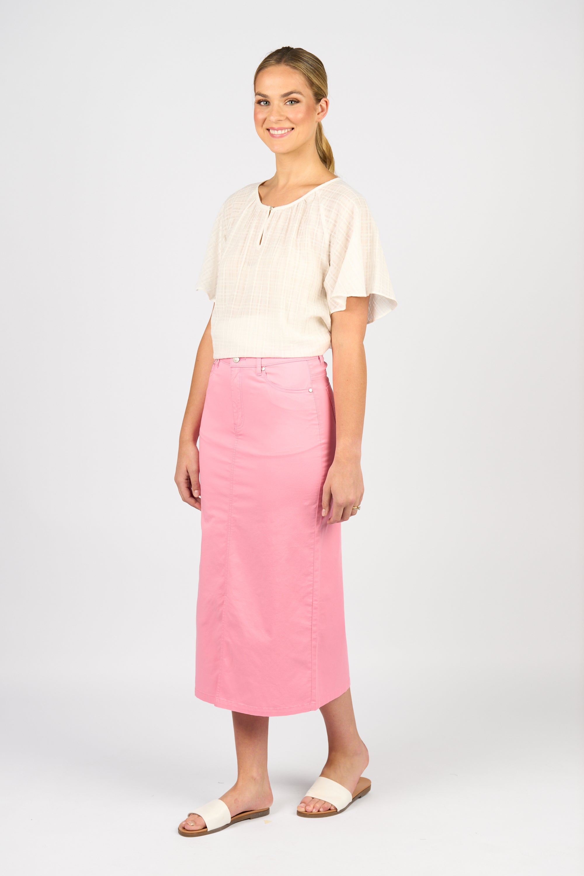 A woman smiles, wearing a short-sleeved light beige blouse, the Vassalli Mid Length Skirt - Jam with a high waist and front split, and white slide sandals, set against a plain white background.