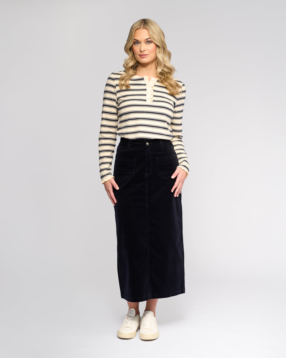 A woman with long blonde hair faces forward, wearing a cream and black striped long-sleeve top, the Vassalli Lane Cord Skirt - Midnight, and white sneakers against a plain light background.