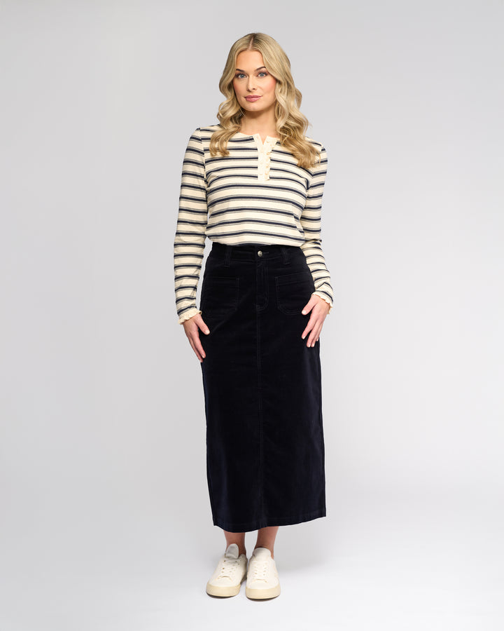 A woman with long blonde hair faces forward, wearing a cream and black striped long-sleeve top, the Vassalli Lane Cord Skirt - Midnight, and white sneakers against a plain light background.