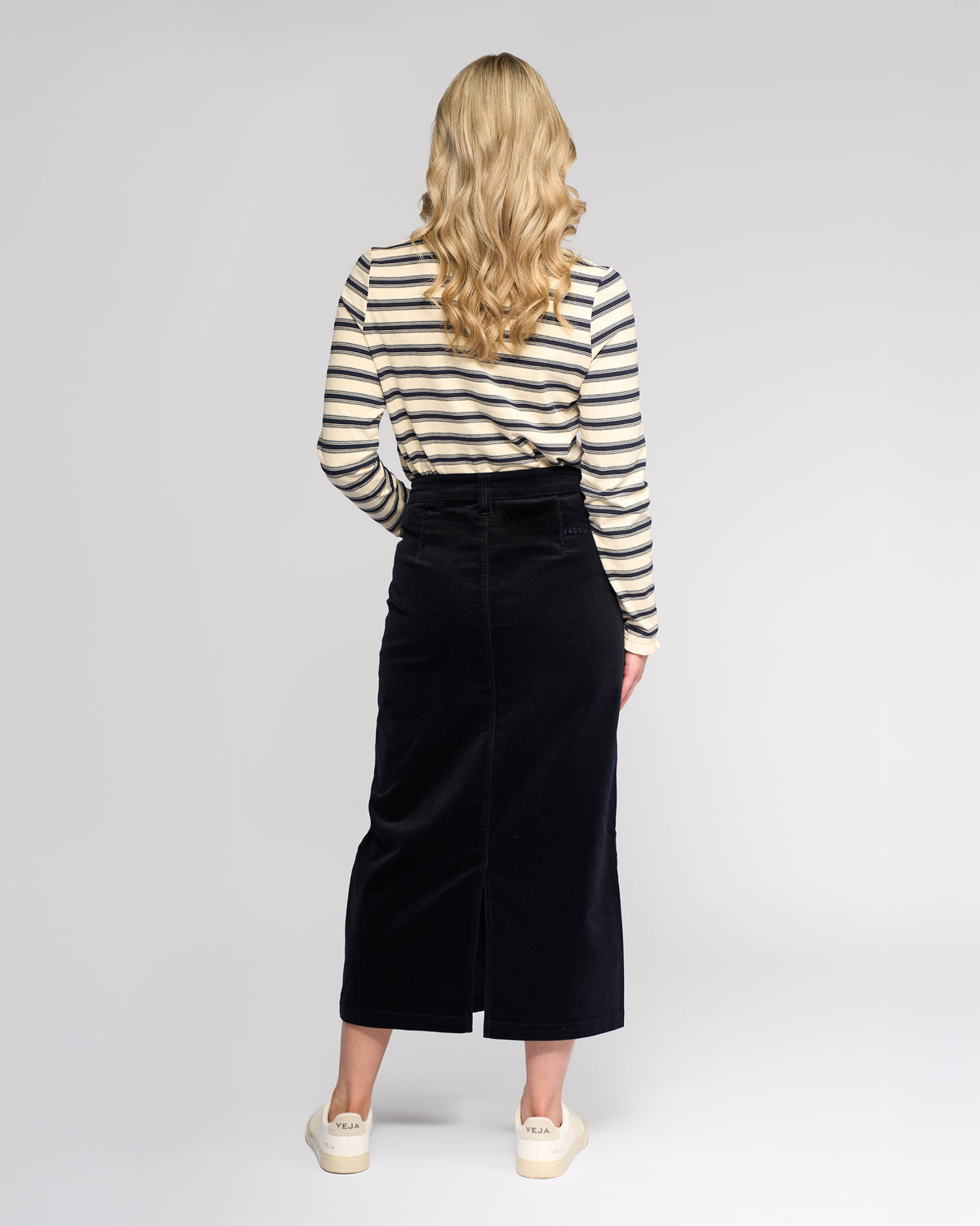 A woman with long wavy blonde hair stands facing away, wearing a cream and navy striped top, cream platform shoes, and the Vassalli Lane Cord Skirt in Midnight against a plain light background.