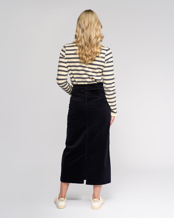 A woman with long wavy blonde hair stands facing away, wearing a cream and navy striped top, cream platform shoes, and the Vassalli Lane Cord Skirt in Midnight against a plain light background.