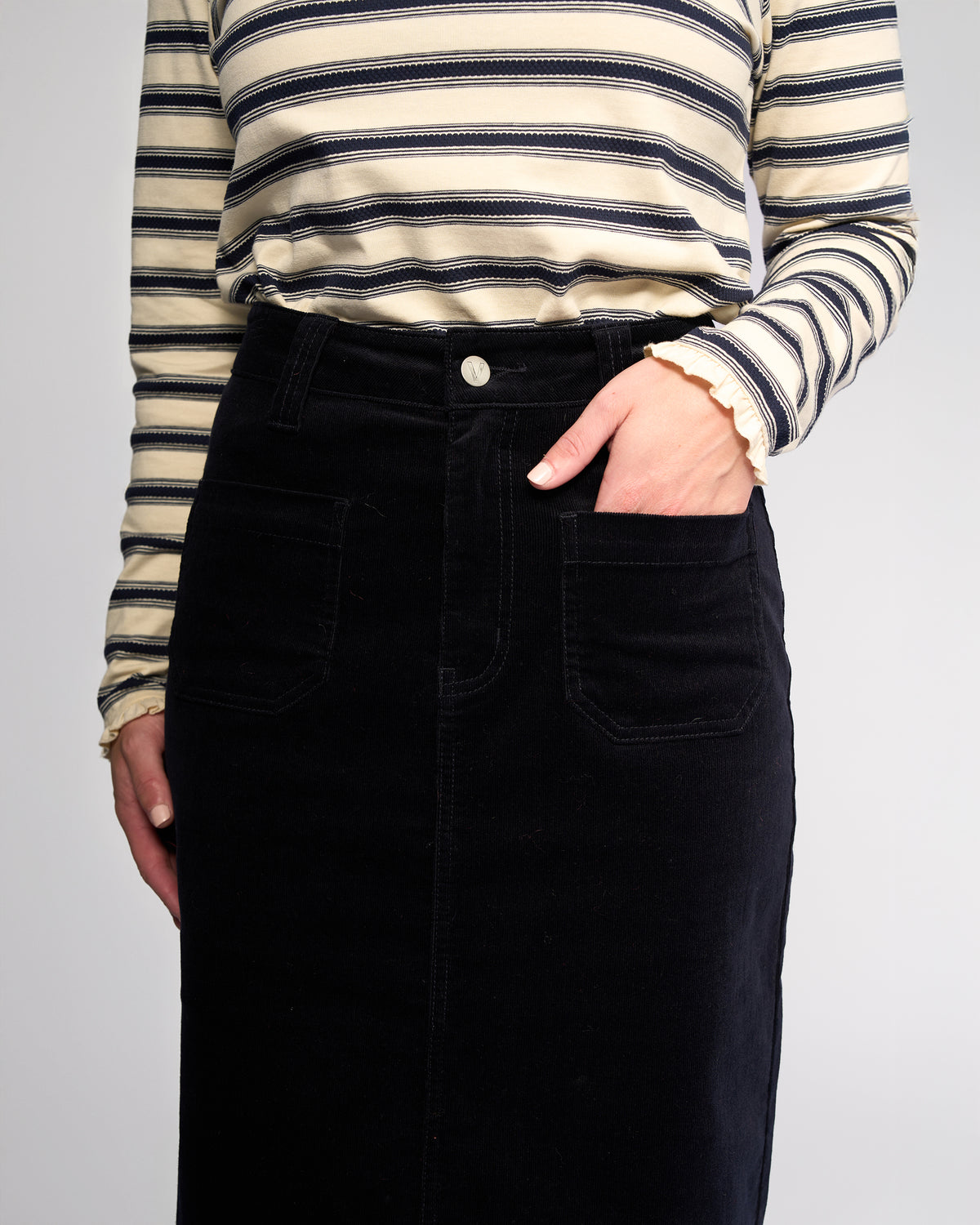 A person wears a cream and navy striped long-sleeve top tucked into the Vassalli Lane Cord Skirt in Midnight, one hand casually in a front pocket. The photo is cropped from shoulders to knees.