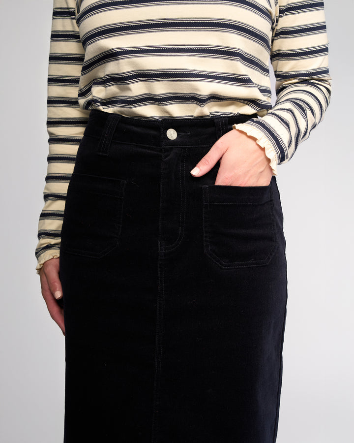 A person wears a cream and navy striped long-sleeve top tucked into the Vassalli Lane Cord Skirt in Midnight, one hand casually in a front pocket. The photo is cropped from shoulders to knees.