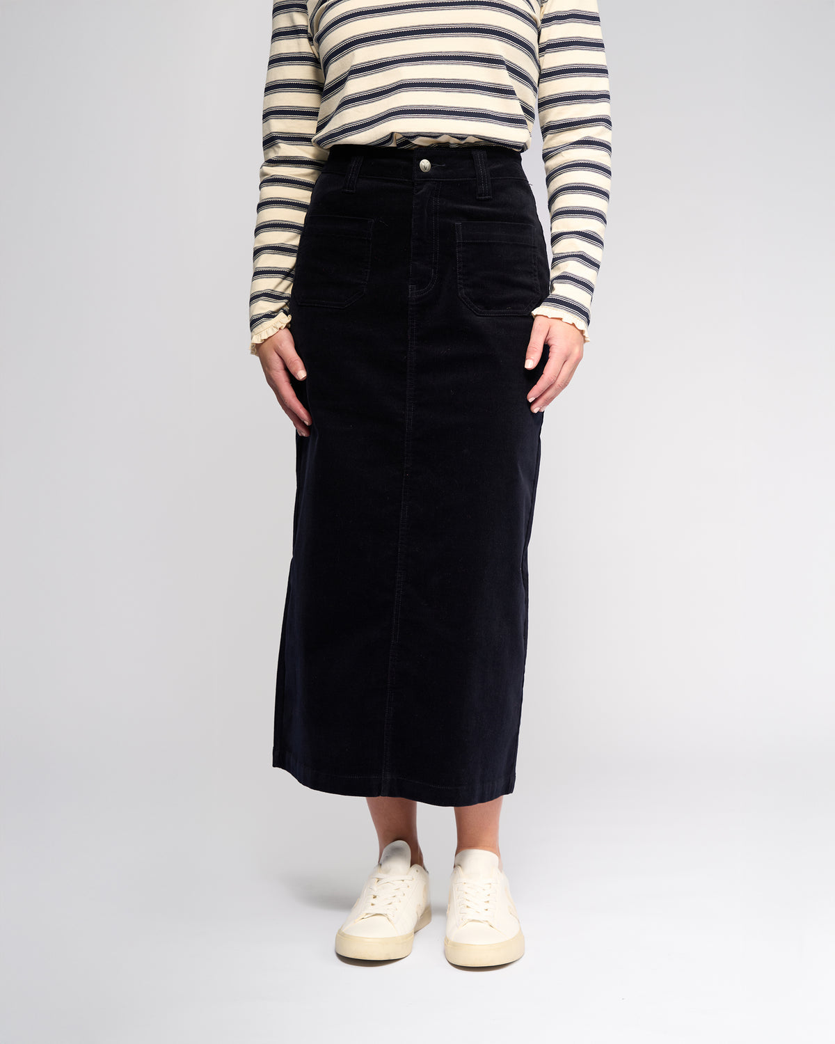 A person models the Vassalli Lane Cord Skirt in Midnight with a cream and navy striped long-sleeve top and white sneakers against a plain light background. Their face is not visible.
