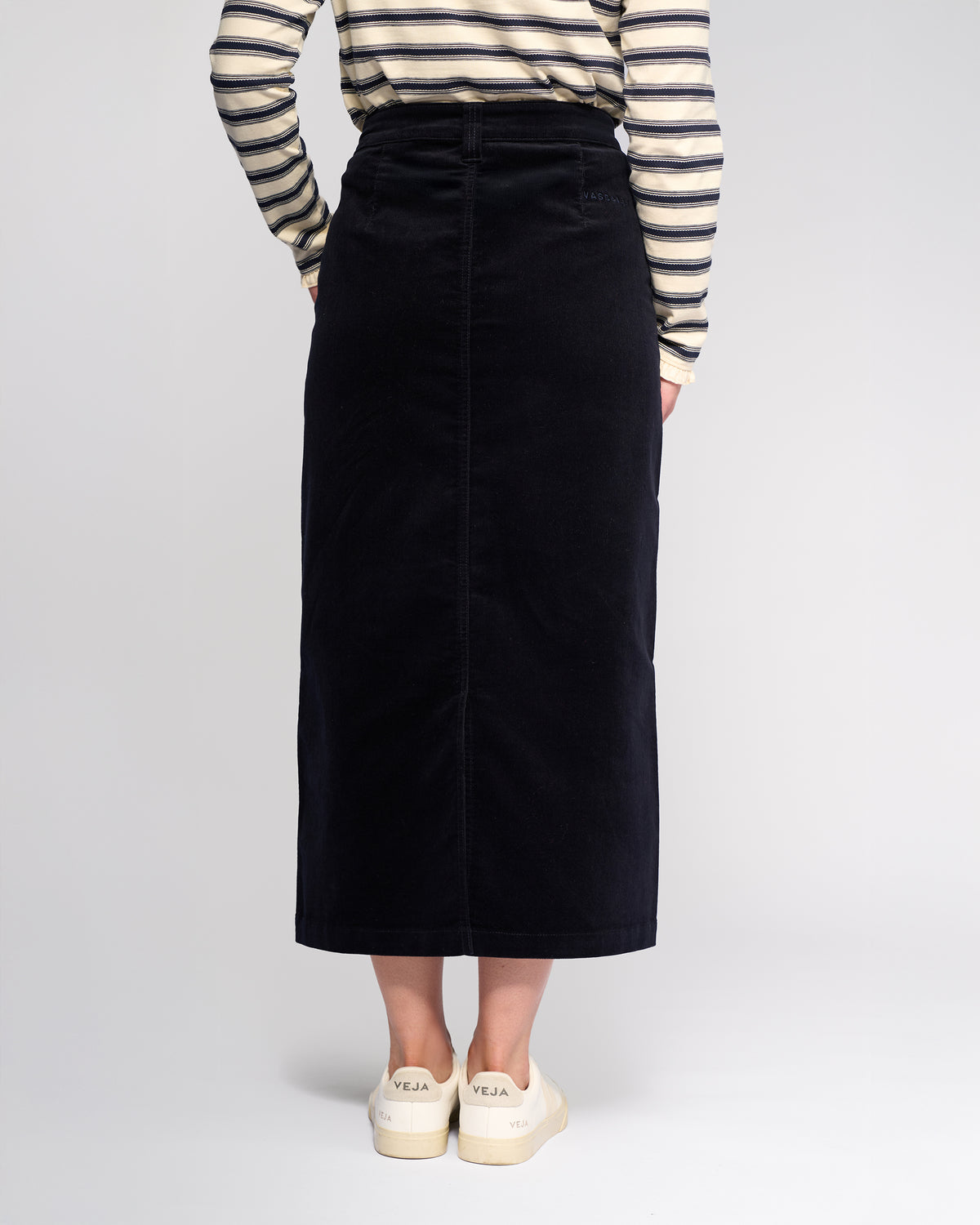 A person stands facing away, wearing the Vassalli Lane Cord Skirt in Midnight, a cream and black striped long-sleeve top, and cream Veja sneakers against a plain light background.