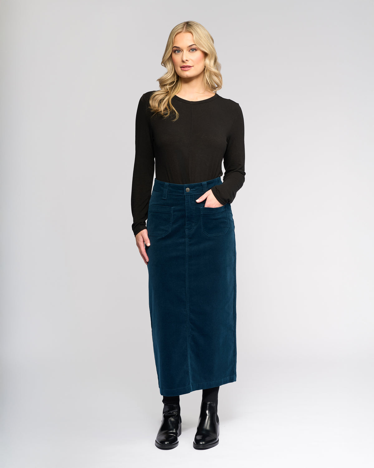 Woman stands against a plain background wearing a long-sleeve black top, Vassalli Lane Cord Skirt in Petrol, black tights, and black ankle boots, with one hand in her pocket.