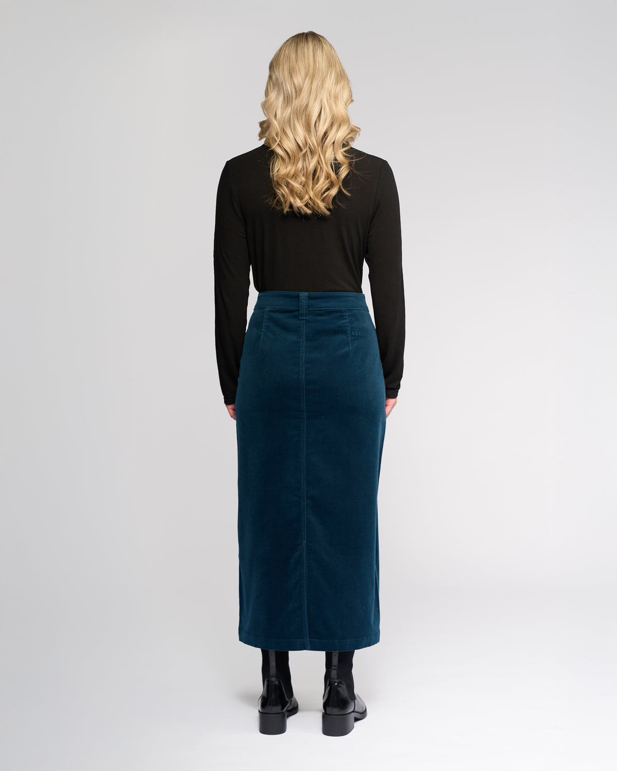 A person with long, wavy blonde hair wears a black long-sleeve top, the Vassalli Lane Cord Skirt in Petrol, and black ankle boots while standing facing away from the camera against a plain light background.