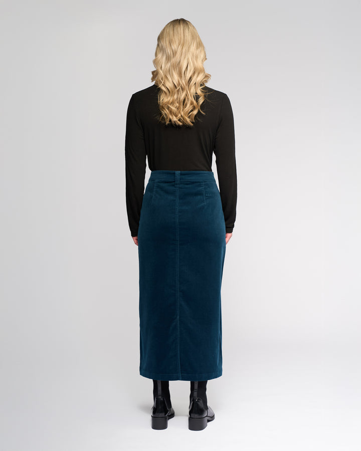 A person with long, wavy blonde hair wears a black long-sleeve top, the Vassalli Lane Cord Skirt in Petrol, and black ankle boots while standing facing away from the camera against a plain light background.