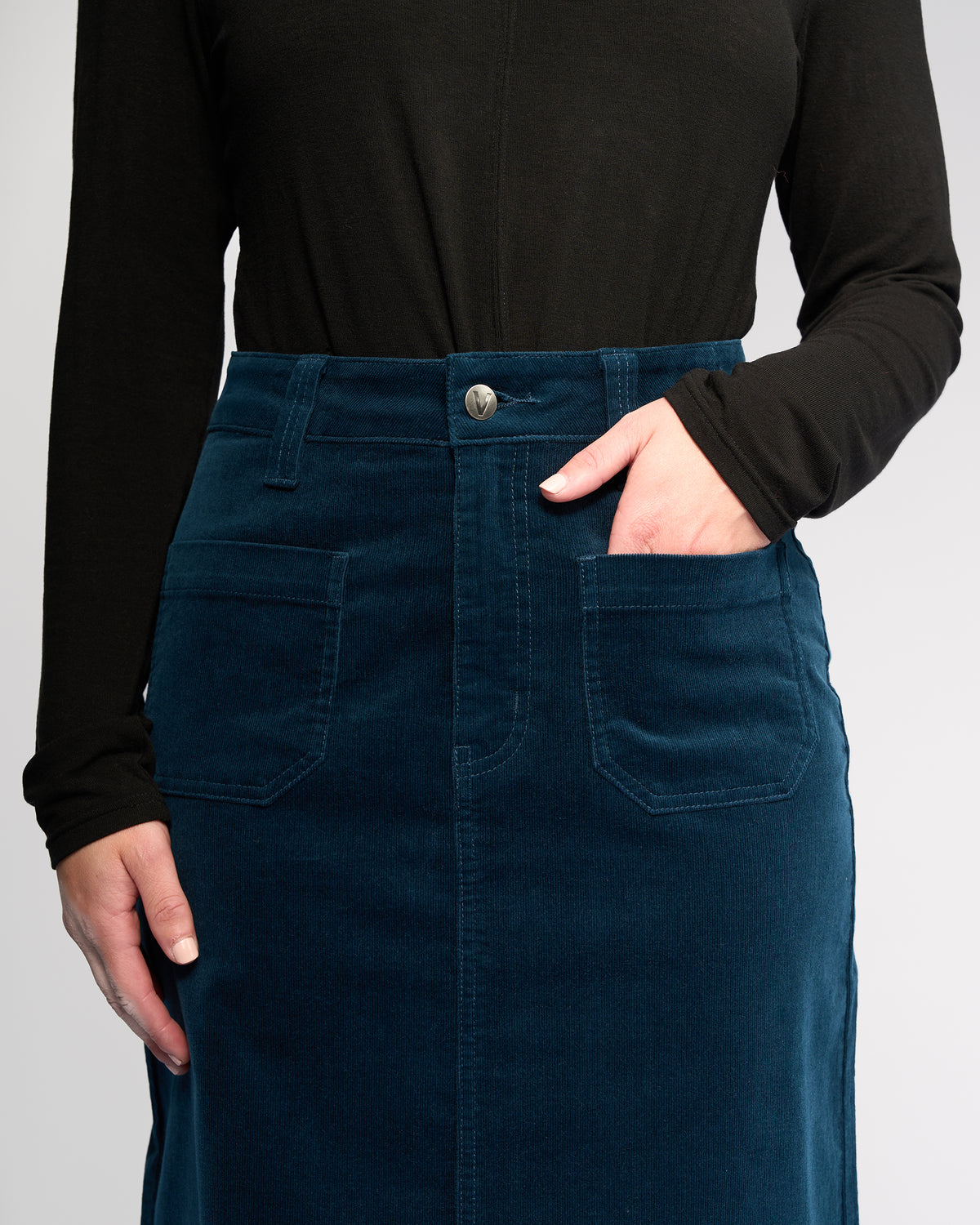A person wearing a long-sleeve black top and the Vassalli Lane Cord Skirt in Petrol—this high-waisted, mid-length stretch corduroy skirt features front pockets, with one hand casually resting in a pocket; shown from shoulders to knees.