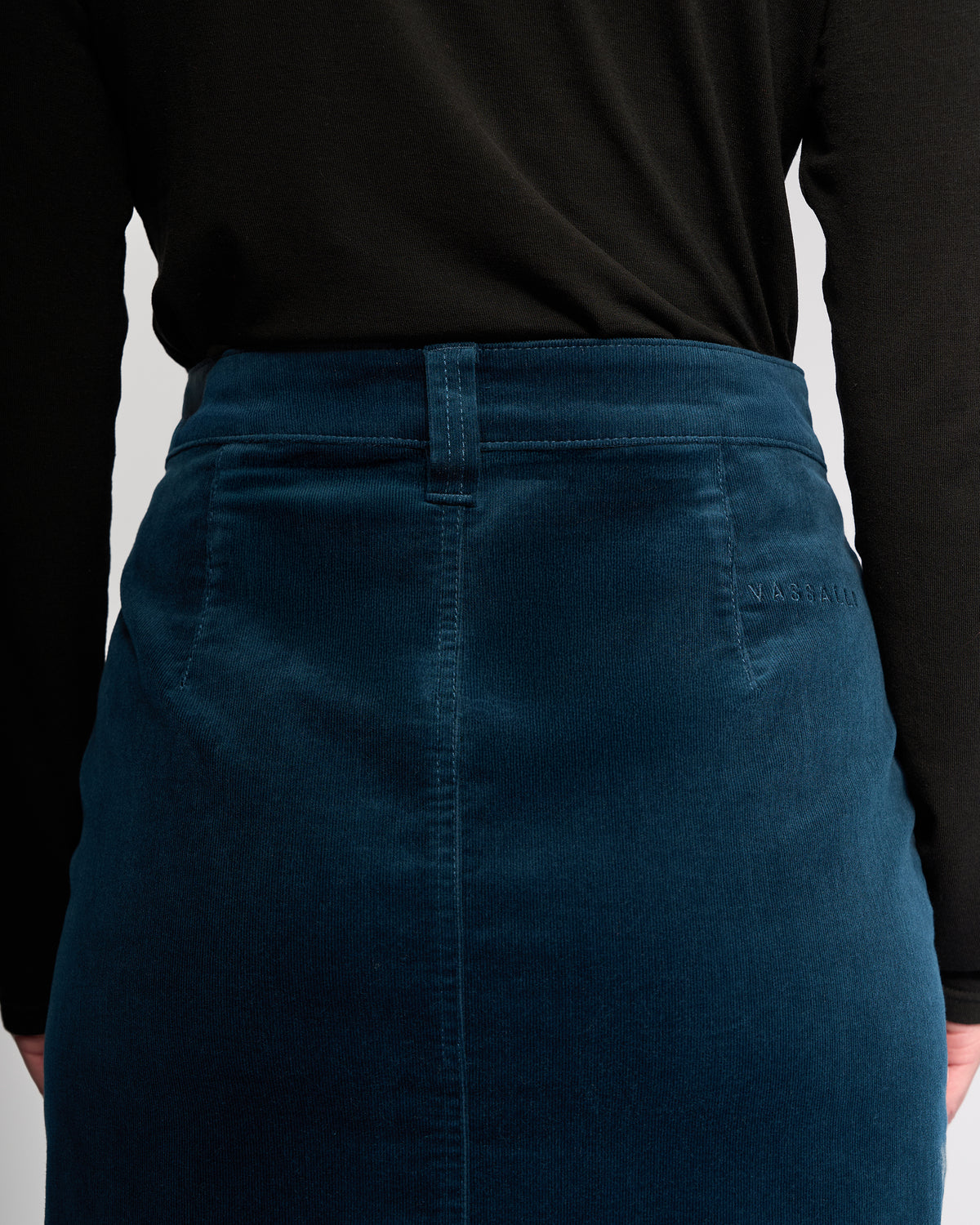 A person seen from the back wears a black long-sleeve top and the Vassalli Lane Cord Skirt in petrol teal stretch corduroy, featuring NASSAU embroidered near the waistband.