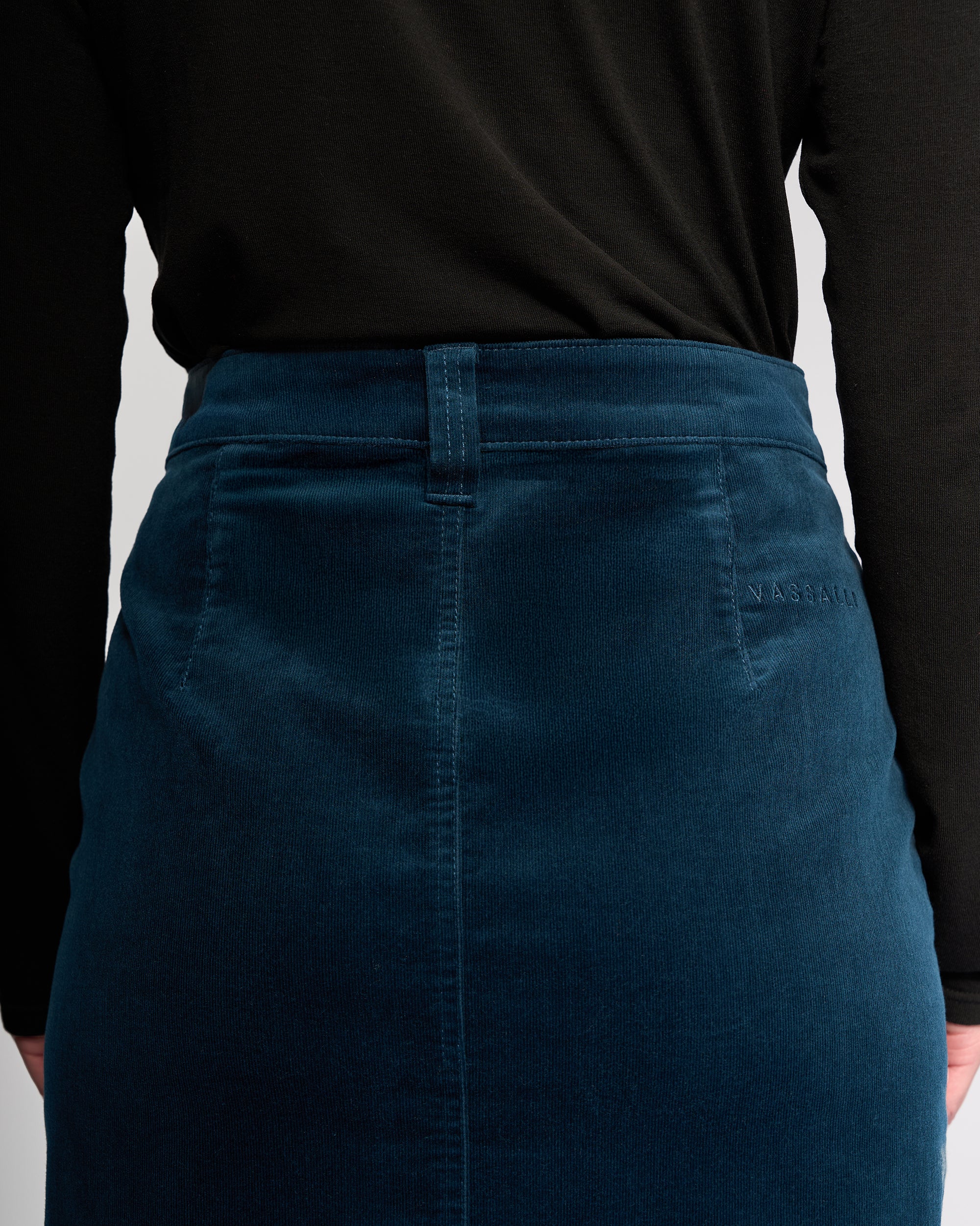 A person seen from the back wears a black long-sleeve top and the Vassalli Lane Cord Skirt in petrol teal stretch corduroy, featuring NASSAU embroidered near the waistband.
