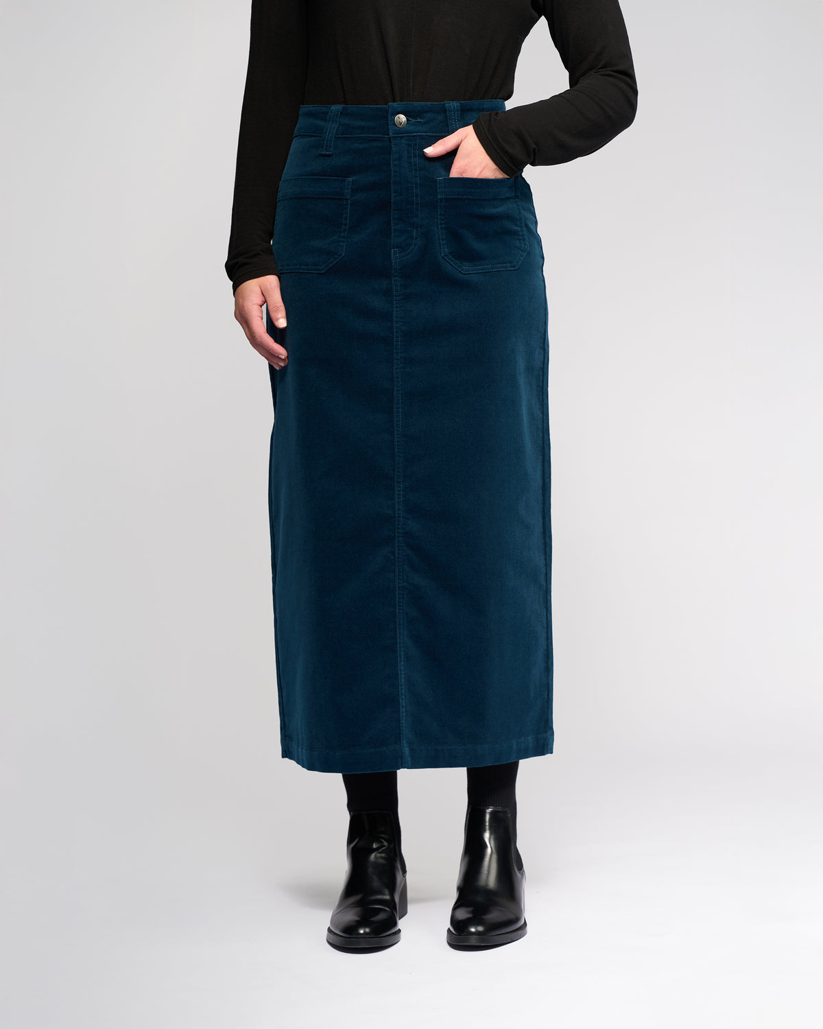 A person wearing a black long-sleeve top, the Vassalli Lane Cord Skirt in Petrol with front pockets, and black ankle boots stands against a plain light background.