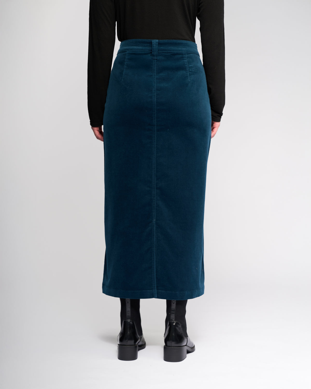 A person faces away from the camera wearing the Vassalli Lane Cord Skirt in petrol, paired with a black long-sleeve top and black ankle boots, set against a plain light background.
