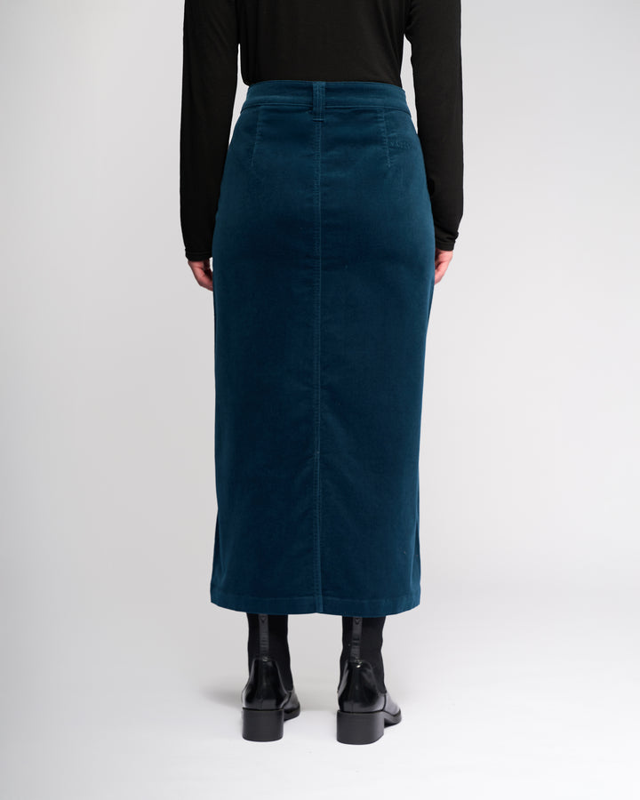 A person faces away from the camera wearing the Vassalli Lane Cord Skirt in petrol, paired with a black long-sleeve top and black ankle boots, set against a plain light background.