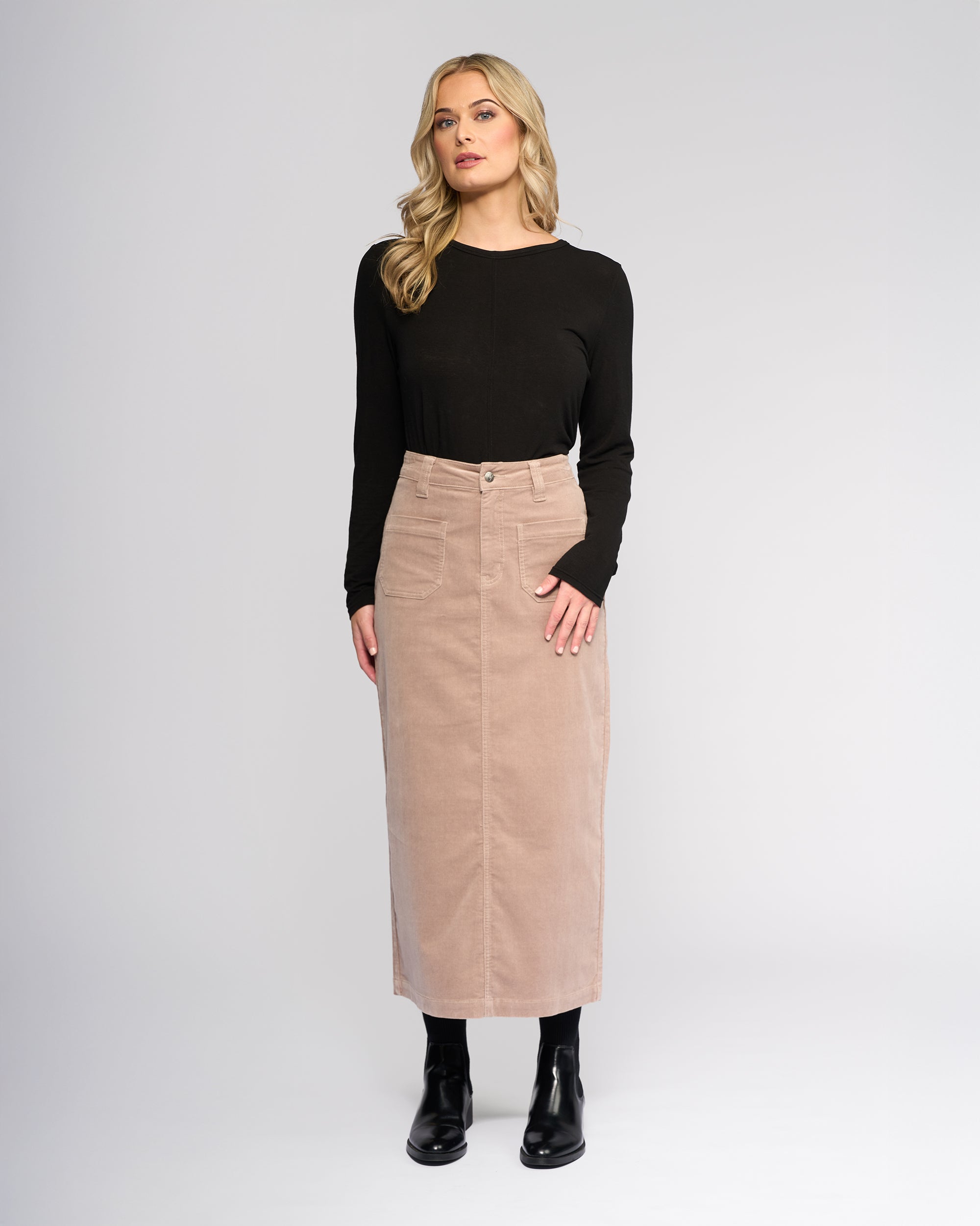A woman with long blonde hair wears a black top, black boots, and the Vassalli Lane Cord Skirt in Twig—a high-waisted, neutral mid-length stretch corduroy skirt—standing against a plain light background.