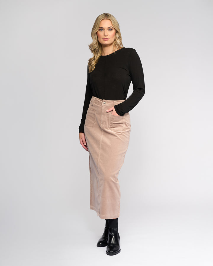 A woman with wavy blonde hair wears a long-sleeve black top, Vassalli Lane Cord Skirt - Twig in a beige high-waisted midi style, and black ankle boots, standing with one hand in her skirt pocket against a plain light background.