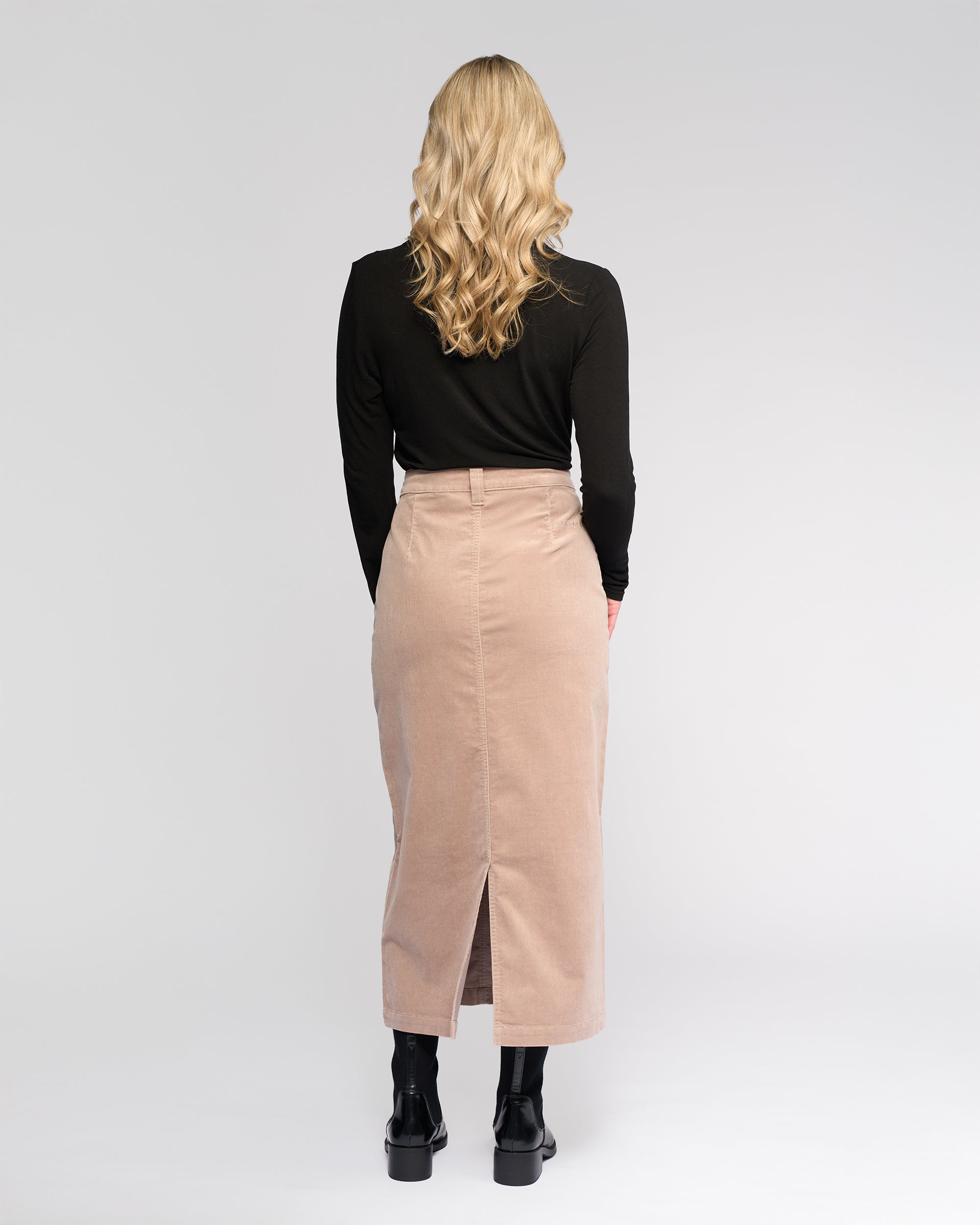 A woman with long blonde hair stands facing away, wearing a black long-sleeve top, black ankle boots, and the Vassalli Lane Cord Skirt in Twig—a winter-ready stretch corduroy skirt with a back slit—against a plain light background.
