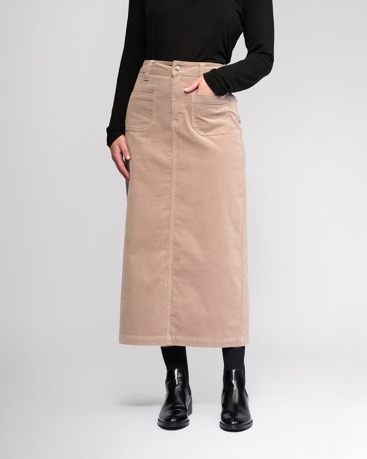 Wearing the Vassalli Lane Cord Skirt - Twig, a beige mid-length corduroy skirt with front pockets, paired with a black long-sleeve top, black tights, and ankle boots—ideal for a winter-ready look against a light background.