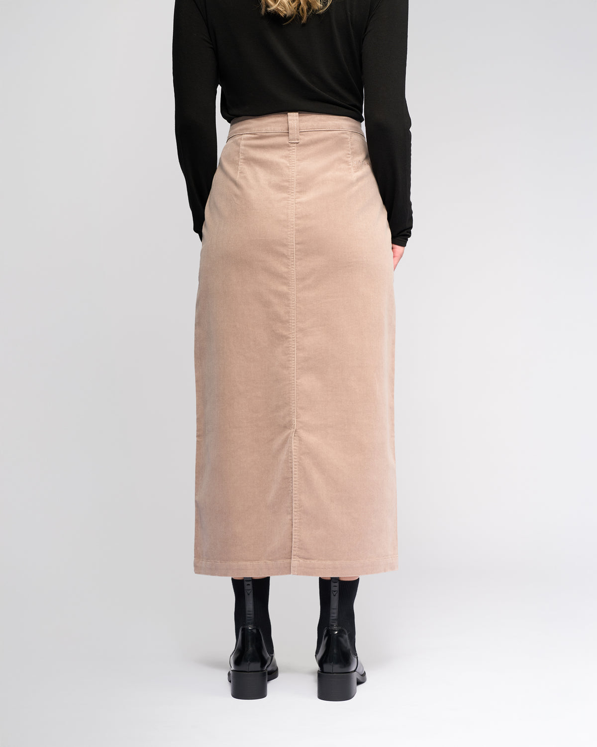 A person stands facing away in a black long-sleeve top, black tights, and boots, wearing the Vassalli Lane Cord Skirt in Twig against a plain light background.
