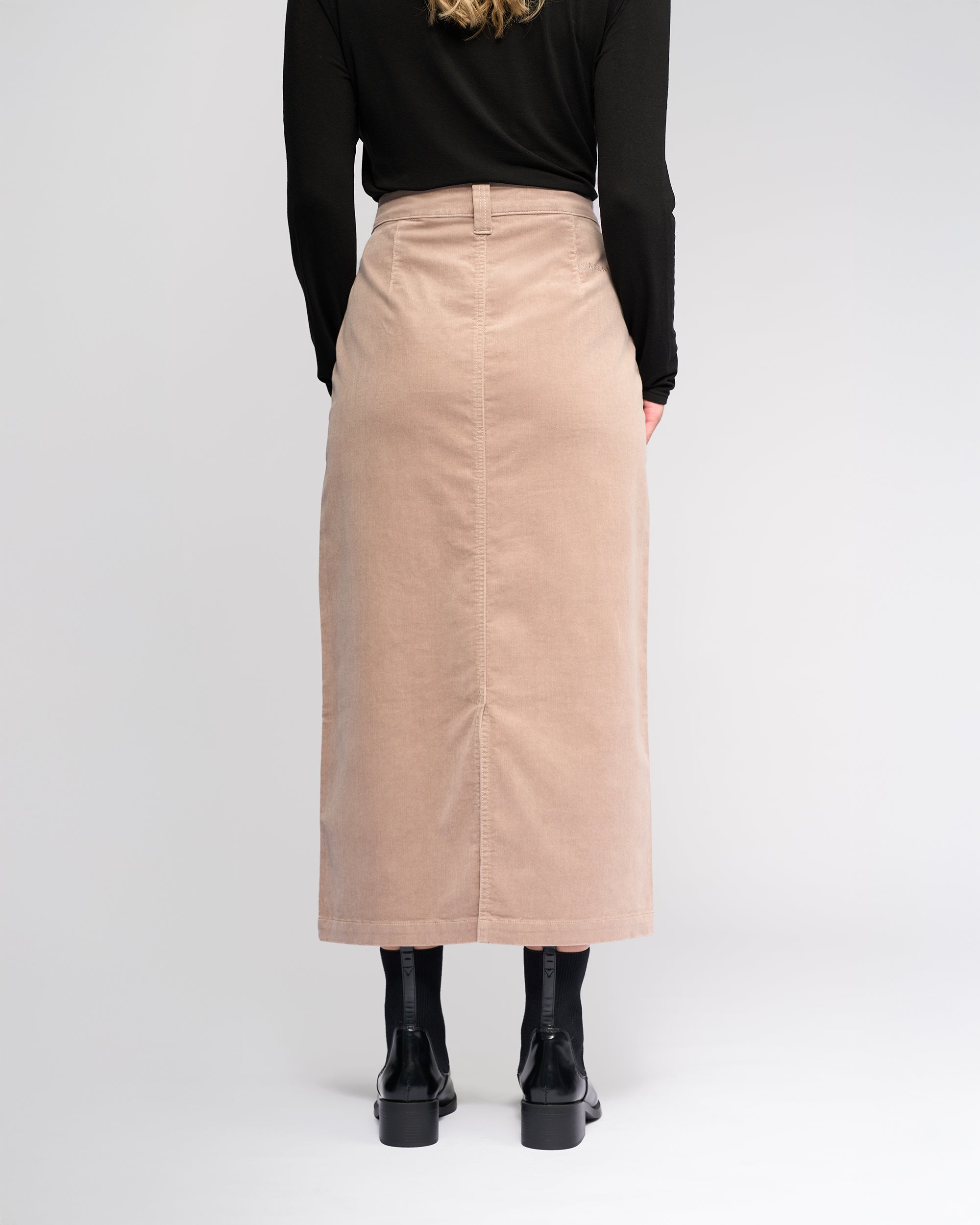A person stands facing away in a black long-sleeve top, black tights, and boots, wearing the Vassalli Lane Cord Skirt in Twig against a plain light background.