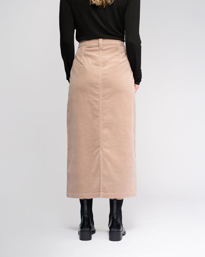 A person stands facing away in a black long-sleeve top, black tights, and boots, wearing the Vassalli Lane Cord Skirt in Twig against a plain light background.