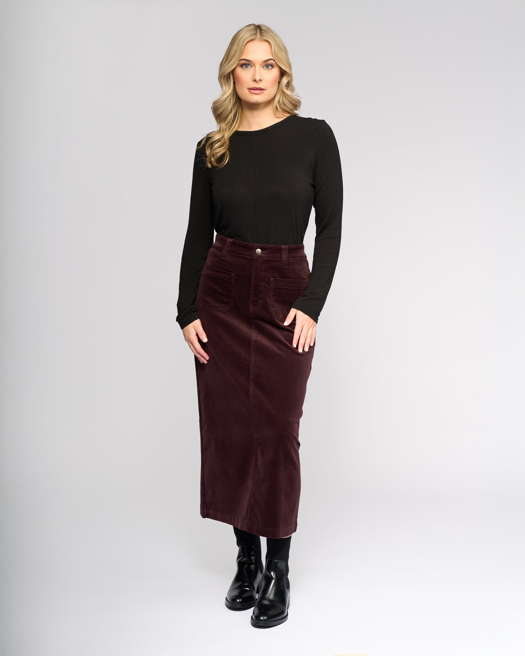 A woman faces forward in a long-sleeve black top, black boots, and the Vassalli Lane Cord Skirt in Merlot, standing against a plain light gray background.