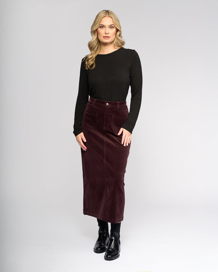 A woman faces forward in a long-sleeve black top, black boots, and the Vassalli Lane Cord Skirt in Merlot, standing against a plain light gray background.