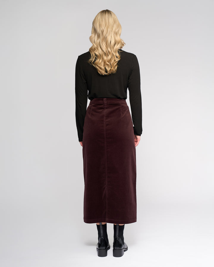 A woman with long, wavy blonde hair faces away, wearing a black long-sleeve top, black shoes, and the Vassalli Lane Cord Skirt in Merlot against a plain light gray background.