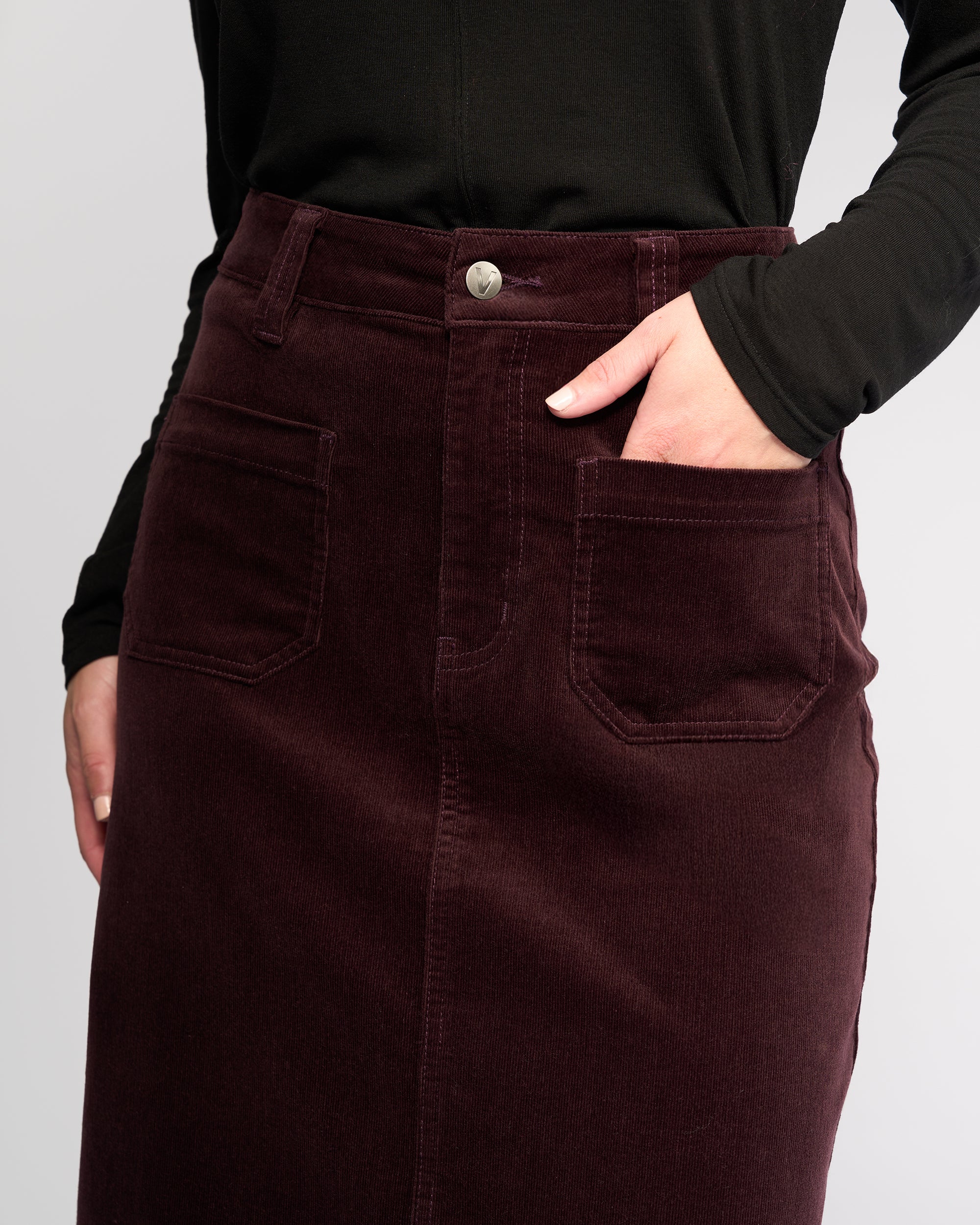 A person wearing a black long-sleeve top and the Vassalli Lane Cord Skirt in Merlot, a maroon red mid-length corduroy skirt with front pockets, stands with one hand in a pocket.