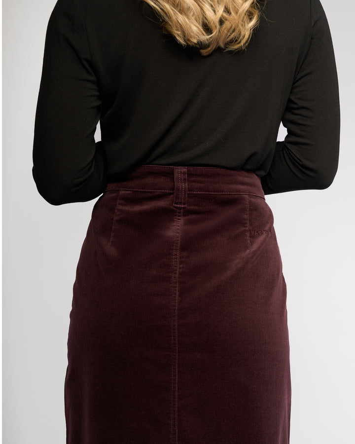 A person with blonde hair, wearing a black long-sleeve top and the Vassalli Lane Cord Skirt in Merlot, is shown from the back against a plain light background.