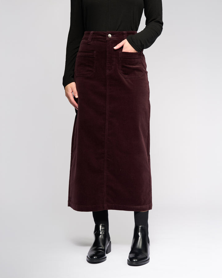 A person wears the Vassalli Lane Cord Skirt in Merlot, a mid-length stretch corduroy skirt with front pockets, paired with a black long-sleeve top, black tights, and ankle boots, standing against a plain light background.