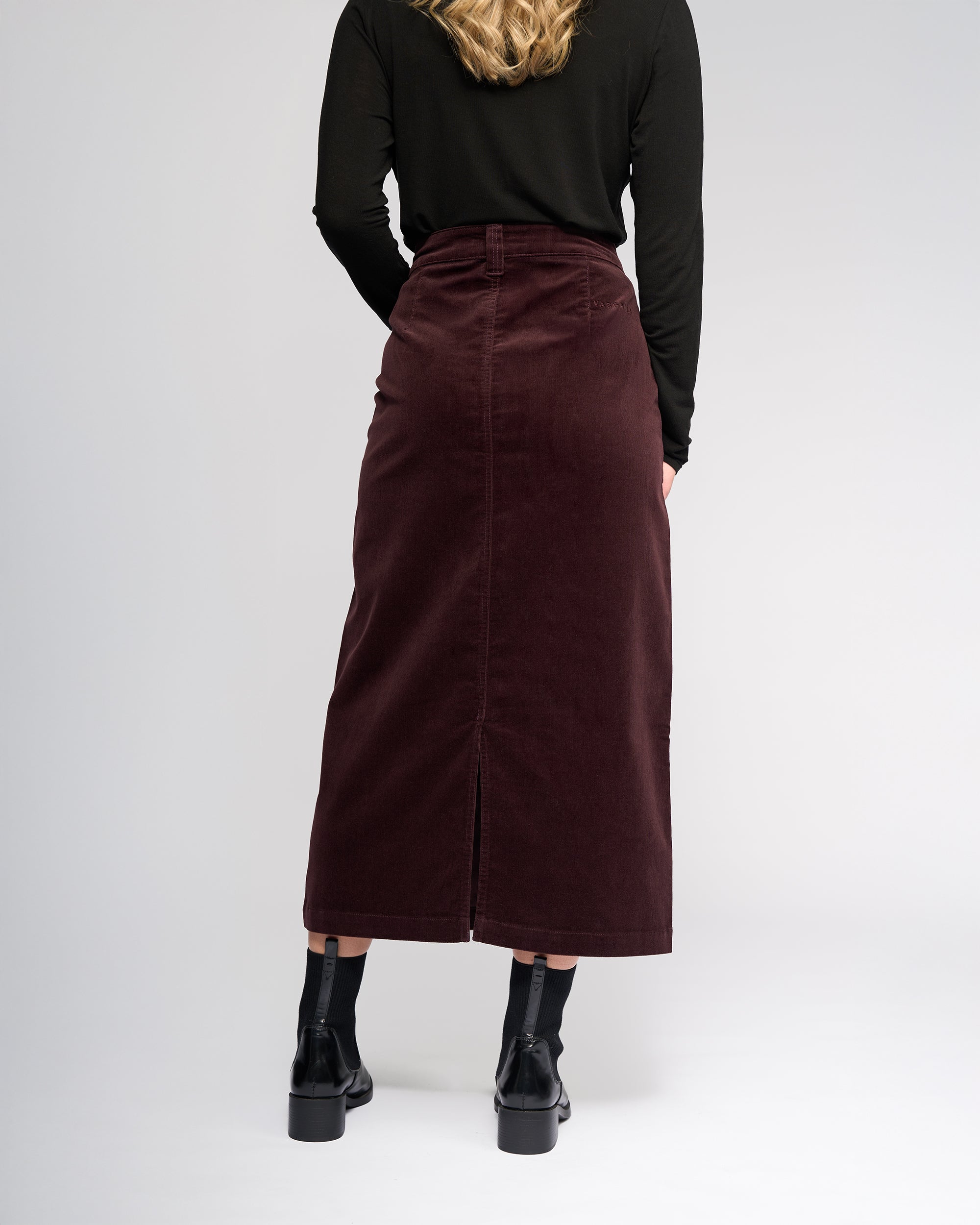 A person with long, wavy blonde hair is seen from behind wearing a black long-sleeve top, Vassalli’s Lane Cord Skirt in Merlot, and black ankle boots against a plain background.